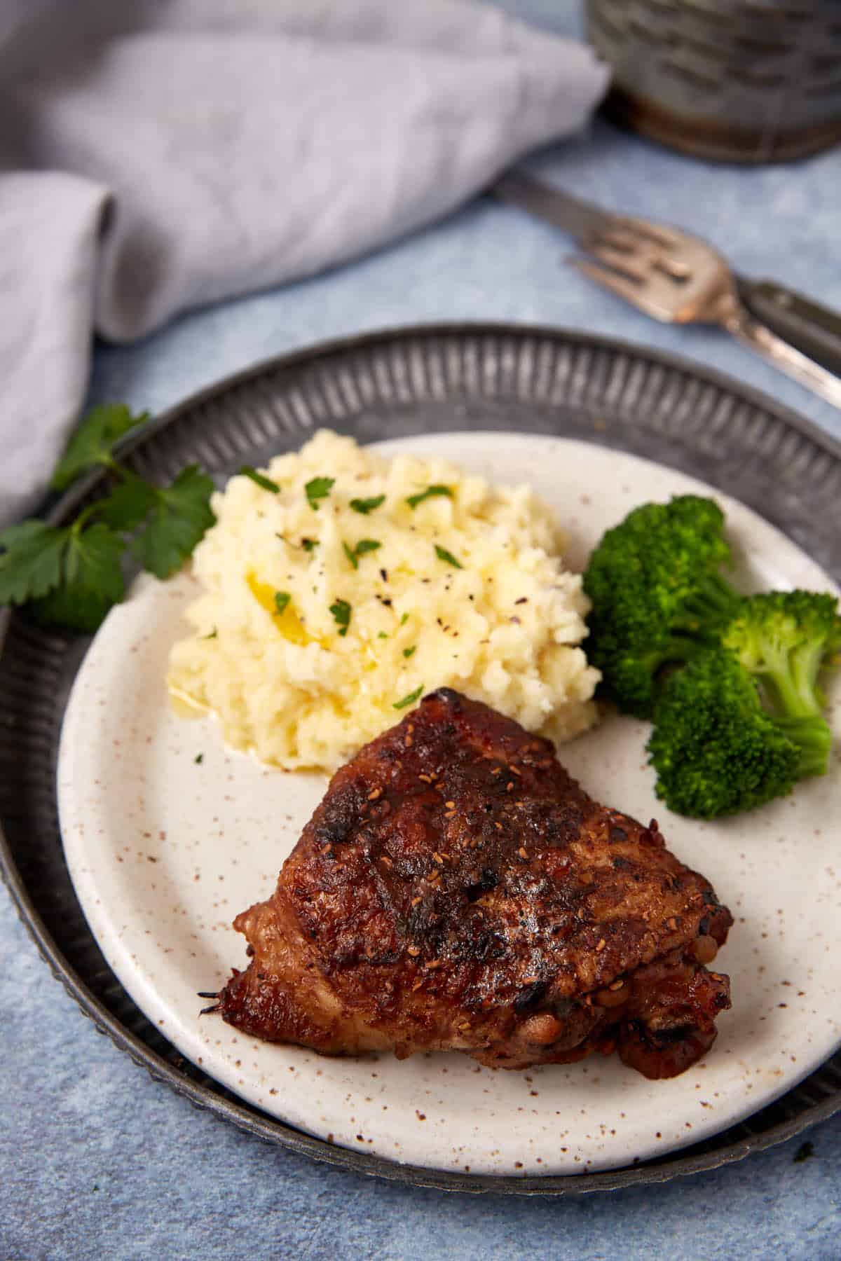 A plate with a piece of seasoned grilled chicken, a serving of mashed potatoes topped with herbs, and steamed broccoli, set on a table with a fork and a gray napkin nearby.