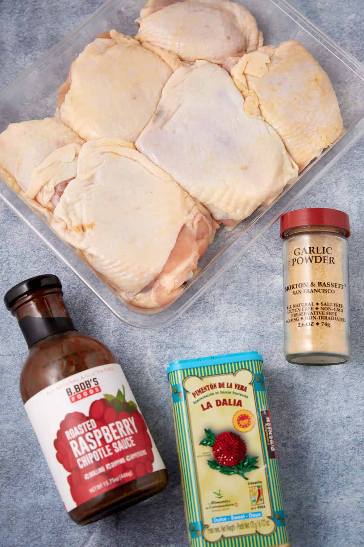 A package of raw chicken thighs sits on a gray surface, surrounded by bottles of raspberry chipotle sauce, smoked paprika, and a jar of garlic powder.
