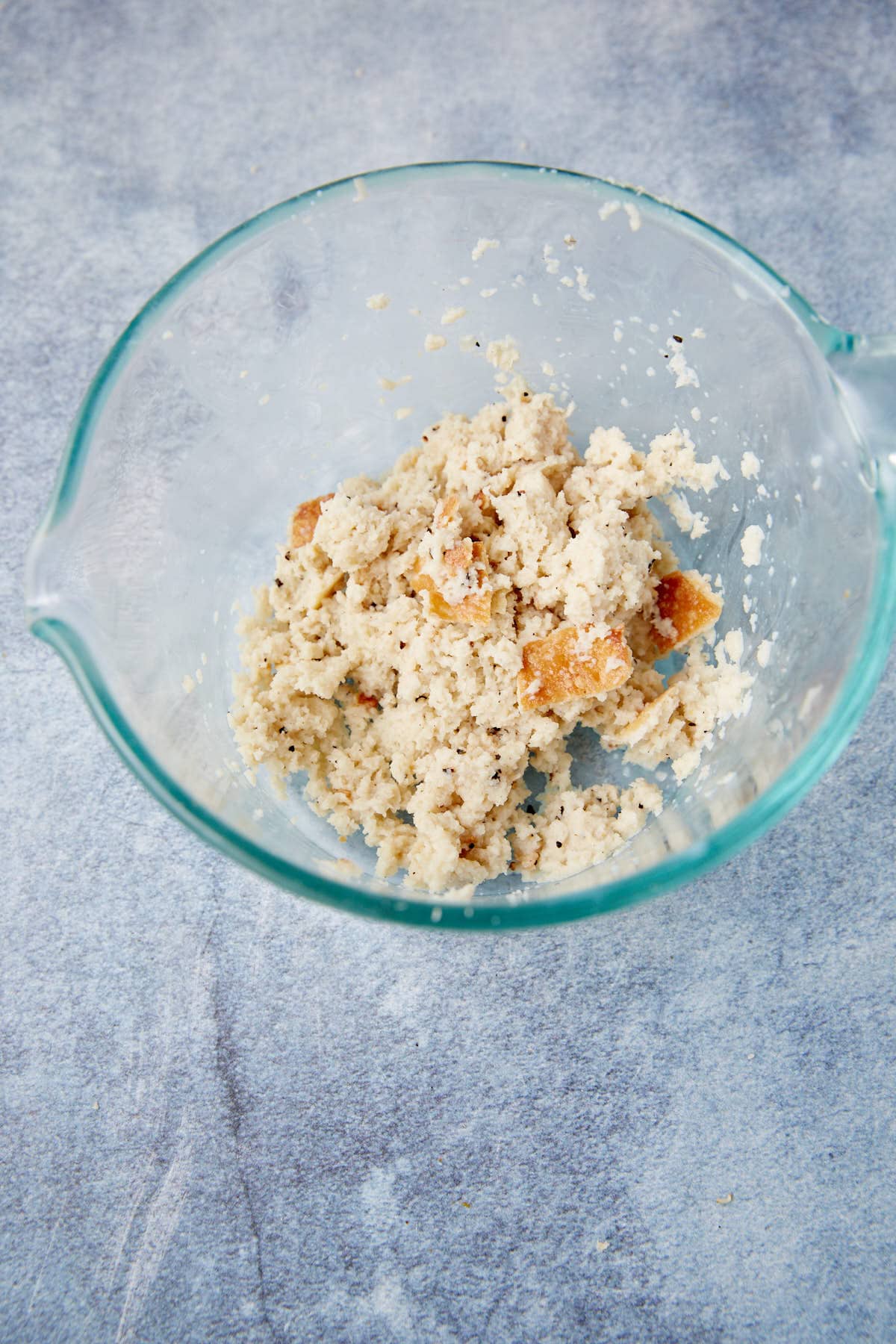 A glass mixing bowl containing a mixture of crumbled bread and liquid on a light gray countertop.