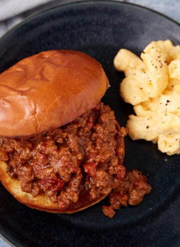 A sloppy joe sandwich on a bun served on a dark plate with a side of creamy macaroni and cheese sprinkled with black pepper.