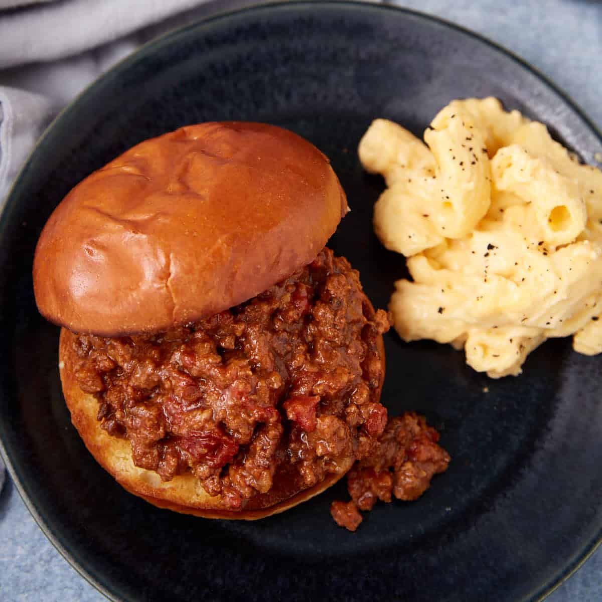 A sloppy joe sandwich on a bun served on a dark plate with a side of creamy macaroni and cheese sprinkled with black pepper.