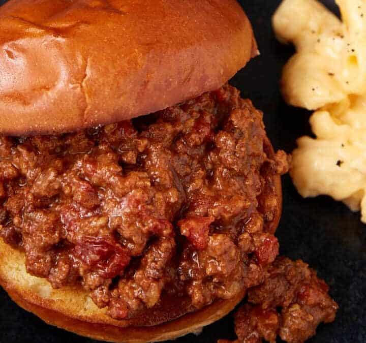 A sloppy joe sandwich with ground beef filling sits on a dark plate next to a serving of creamy macaroni and cheese. A second bun and a pot of meat are visible in the background.