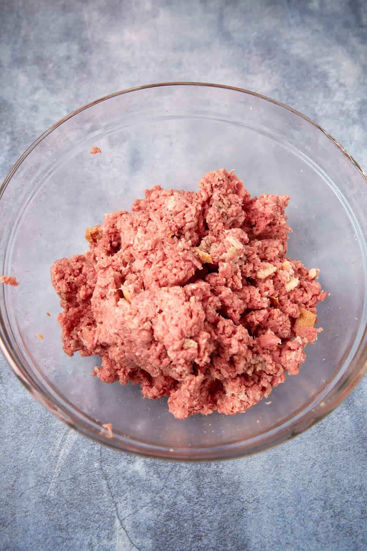 Raw ground beef mixed with seasoning in a clear glass bowl, placed on a blue-gray textured surface.