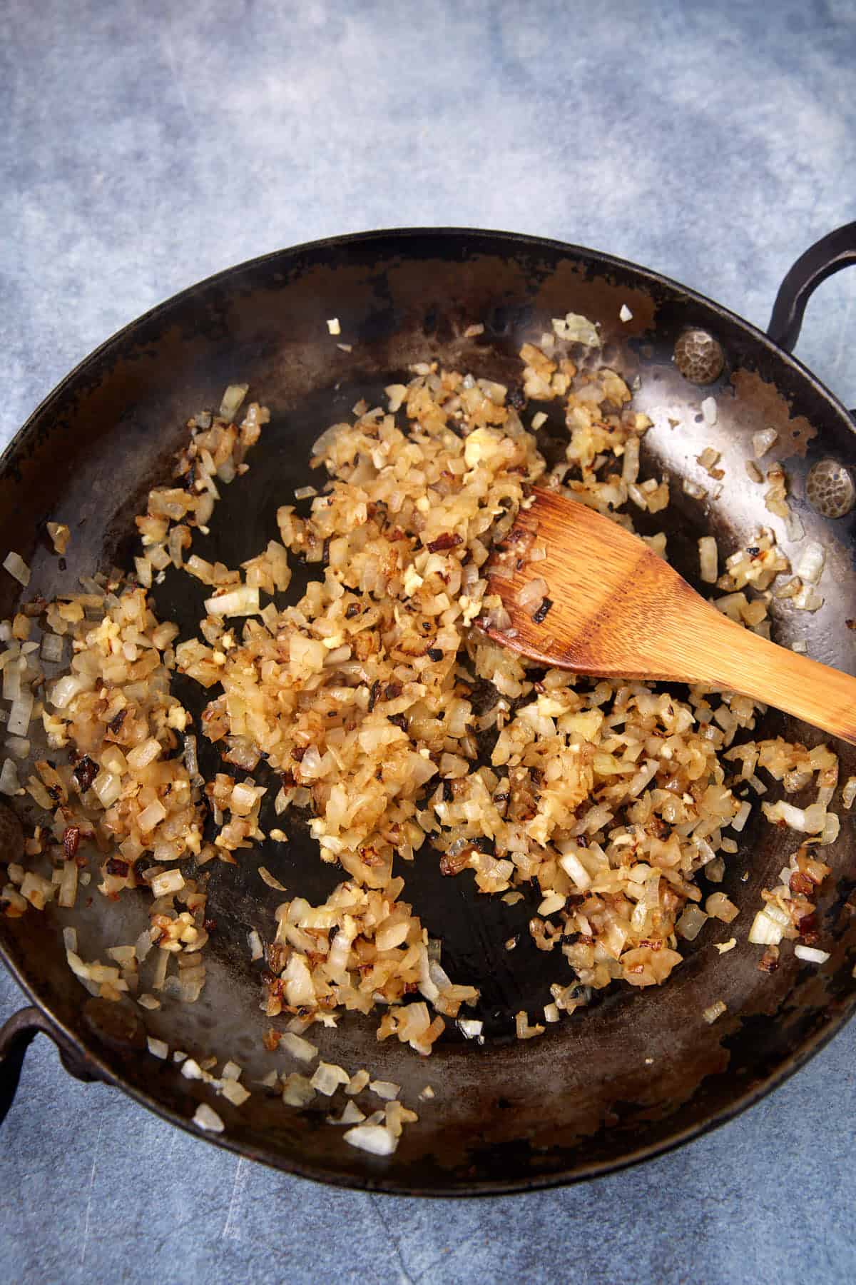 Chopped onions being sautéed in a dark skillet with a wooden spoon, showing golden brown caramelization on a gray textured surface.