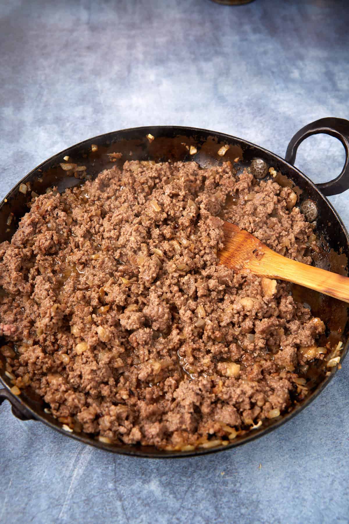 Ground beef and chopped onions are being cooked in a black skillet, with a wooden spoon stirring the mixture. The skillet is on a blue countertop. Steam is visible rising from the hot food.