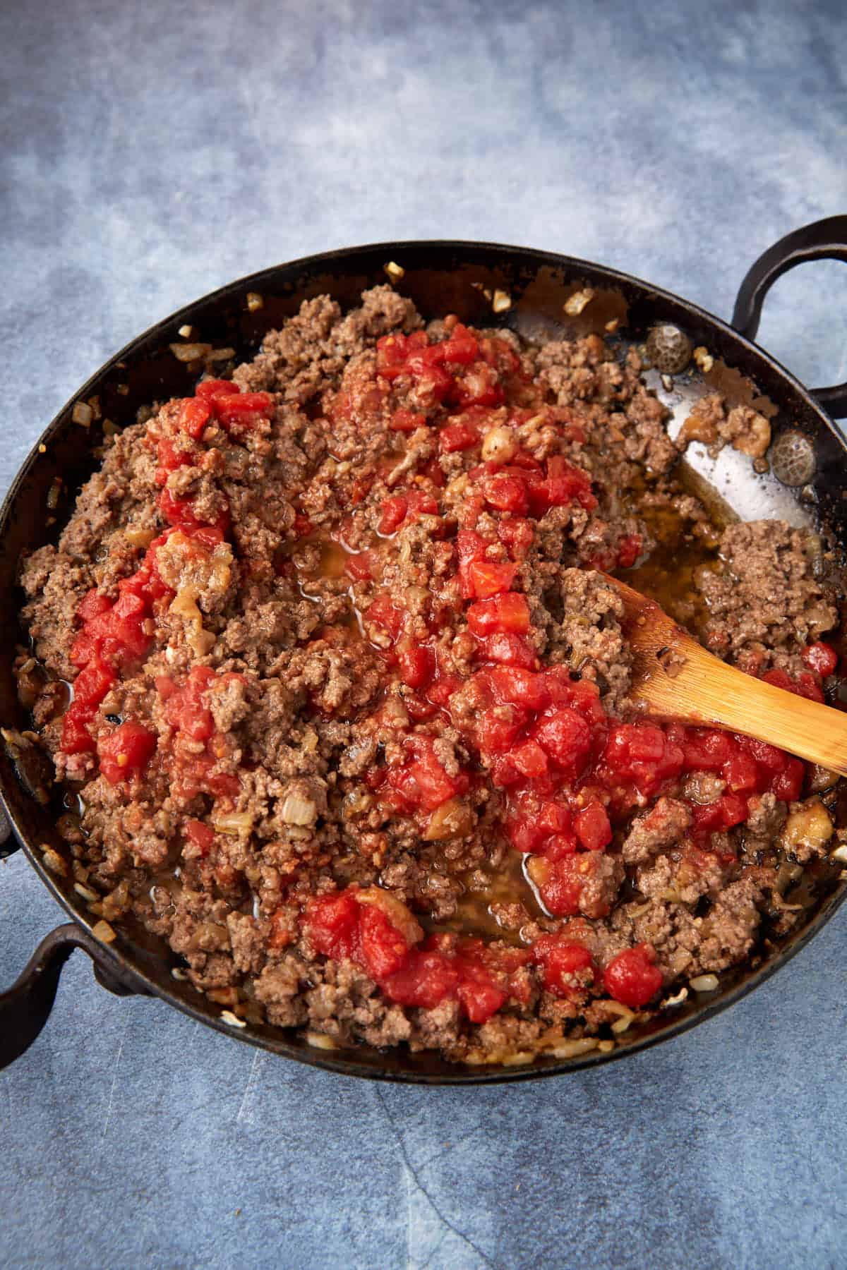 Ground beef, diced tomatoes, and onions are being sautéed in a black skillet with a wooden spoon on a blue surface.