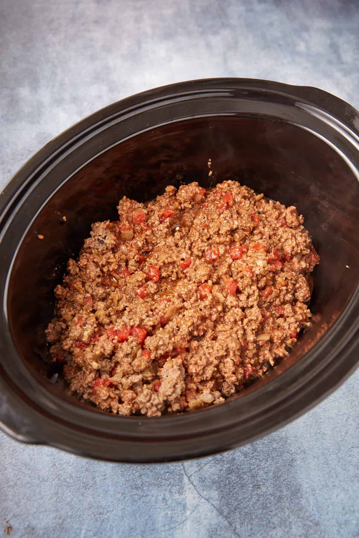 A slow cooker filled with cooked ground beef mixed with diced tomatoes, sitting on a light gray surface.