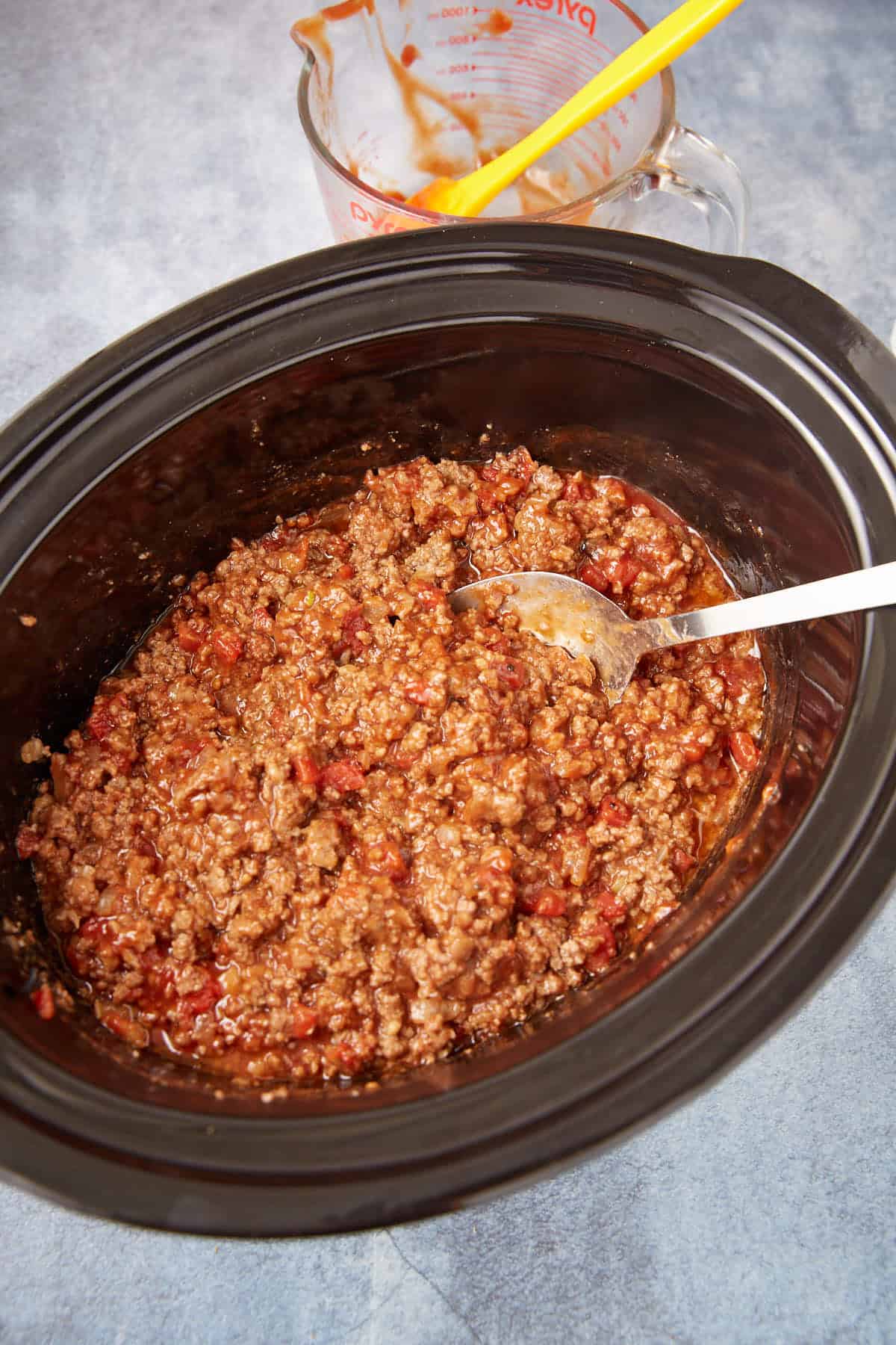 Ground beef and tomato sauce mixture in a black slow cooker with a metal spoon, sitting on a gray surface. In the background, a glass measuring cup with sauce residue and a yellow spatula is visible.