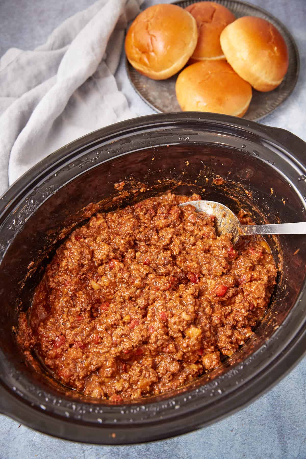 A slow cooker filled with sloppy joe meat mixture with a spoon inside, next to a plate holding four shiny sandwich buns and a light gray napkin.