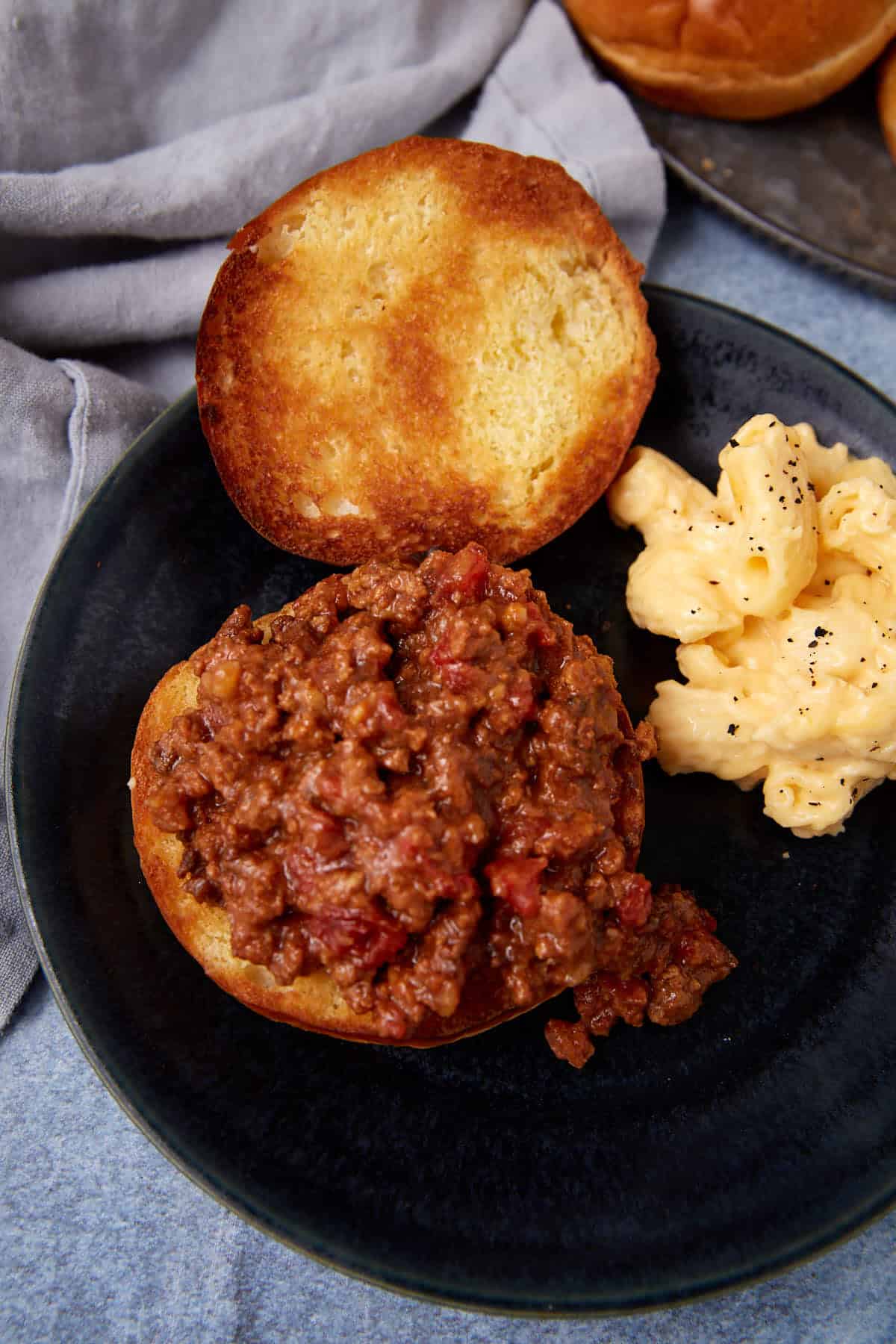 A toasted sandwich bun topped with sloppy joe filling sits on a dark plate next to a serving of creamy macaroni and cheese, with a gray cloth in the background.