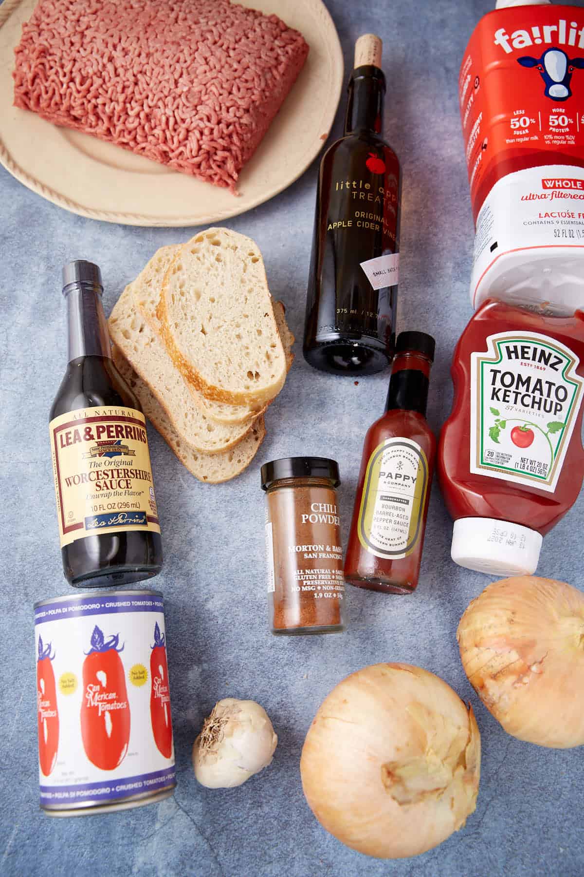 A flat lay of ingredients on a blue surface, including ground beef, sliced bread, onions, garlic, ketchup, Worcestershire sauce, apple cider vinegar, chili powder, canned tomatoes, hot sauce, and a carton of milk.