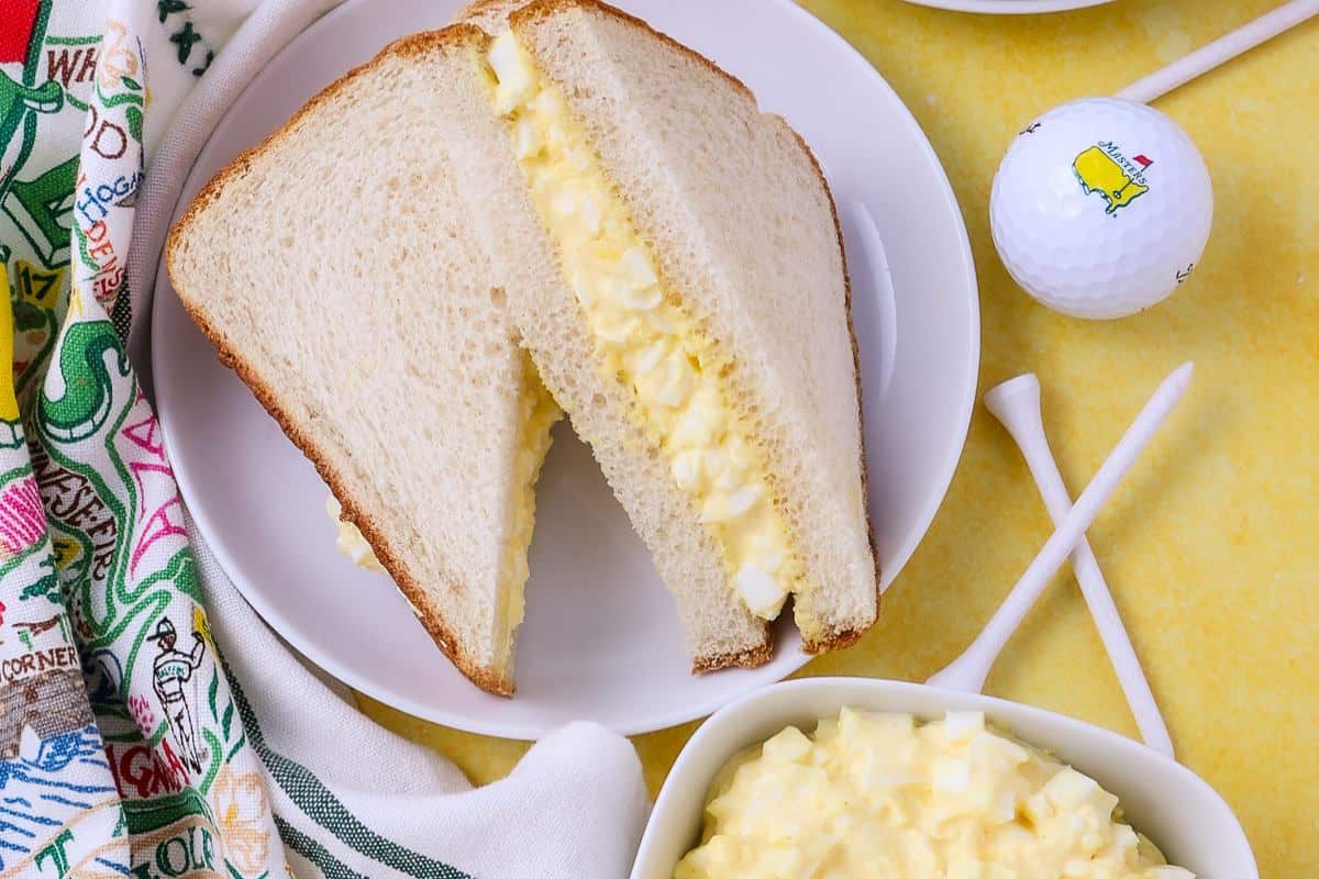A plate with an egg salad sandwich cut in half on white bread, next to a patterned napkin, golf ball, two tees, and a bowl of egg salad on a yellow surface—your Masters party menu isn’t complete without these Southern dishes.