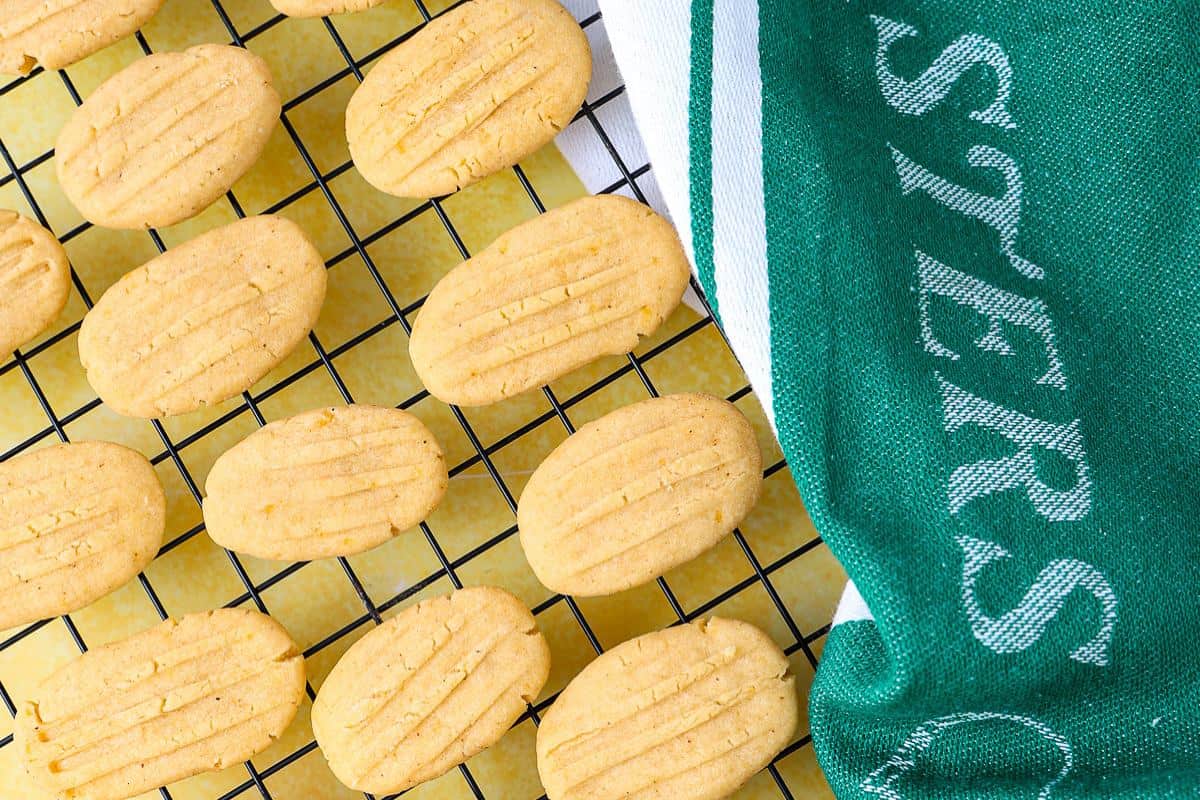 Oval-shaped cookies cool on a black wire rack, next to a green and white kitchen towel with partial white text. Styled on a bright yellow surface, these treats could be among the southern appetizers every Masters party needs.