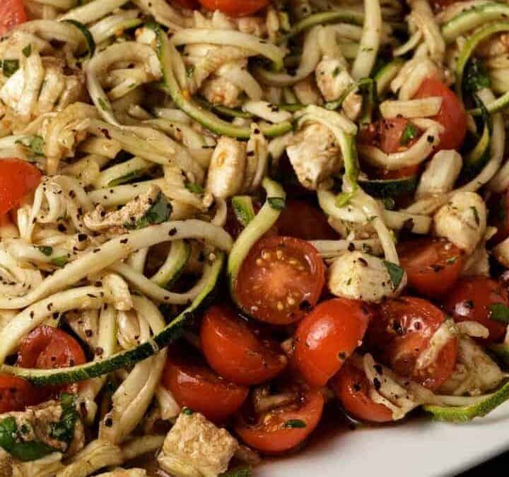 A plate of zucchini noodles mixed with cherry tomatoes, cooked chicken pieces, and herbs, seasoned with black pepper—one of those fresh spring recipes that make winter feel officially over. A wooden spoon is partially visible on the side.