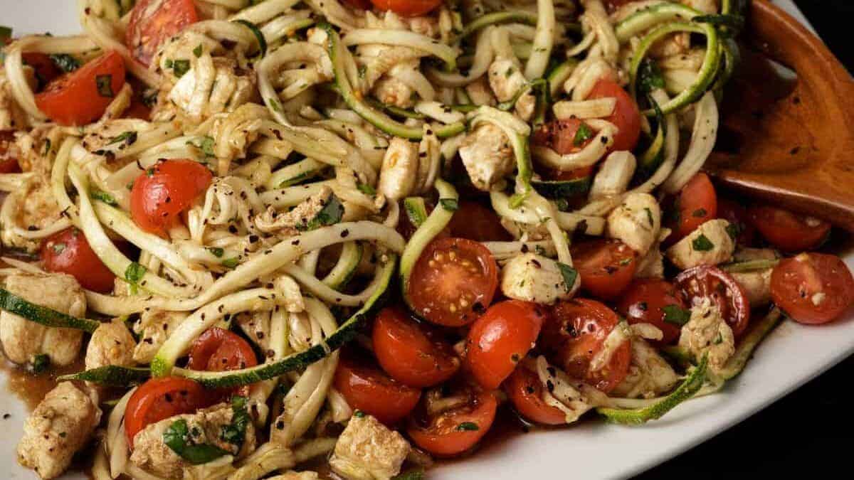A plate of zucchini noodles mixed with cherry tomatoes, cooked chicken pieces, and herbs, seasoned with black pepper—one of those fresh spring recipes that make winter feel officially over. A wooden spoon is partially visible on the side.