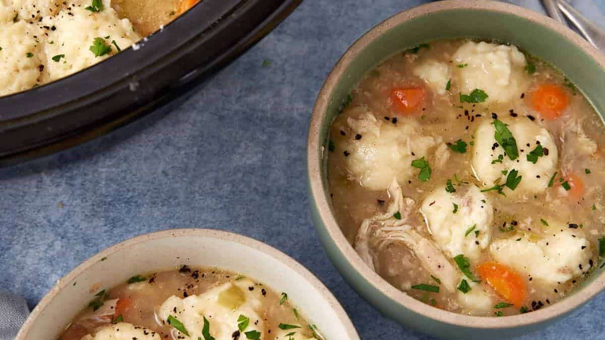 Two bowls and a pot of chicken and dumplings soup with carrots and herbs, set on a blue surface.