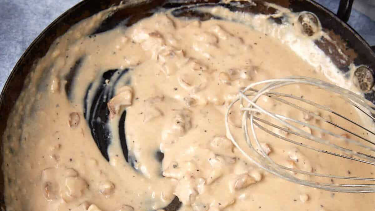 A skillet filled with creamy gravy containing small pieces of sausage, with a metal whisk resting