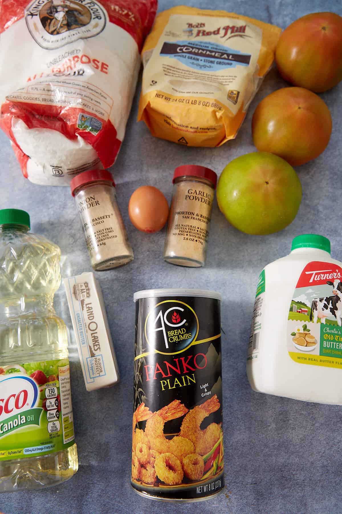 A flat lay of various cooking ingredients for Fried Green Tomatoes: a bag of flour, cornmeal, two grapefruits, garlic powder and onion powder, an egg, butter, canola oil, panko breadcrumbs, and a jug of whole milk.