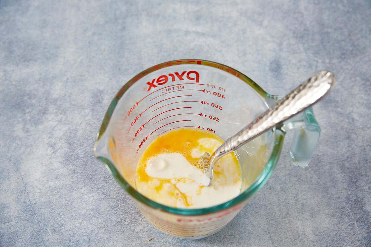 A glass Pyrex measuring cup with cracked eggs and cream being mixed with a silver spoon, perfect for preparing batter for classic dishes like Fried Green Tomatoes, sits on a light blue textured surface.