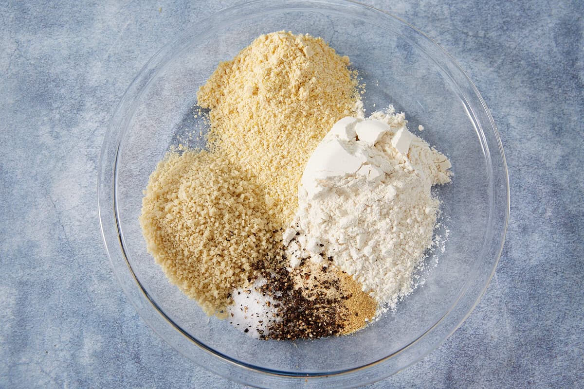A glass bowl containing piles of flour, panko breadcrumbs, crushed cornflakes, salt, ground black pepper, and garlic powder sits on a blue textured surface—perfect for coating classic Fried Green Tomatoes.