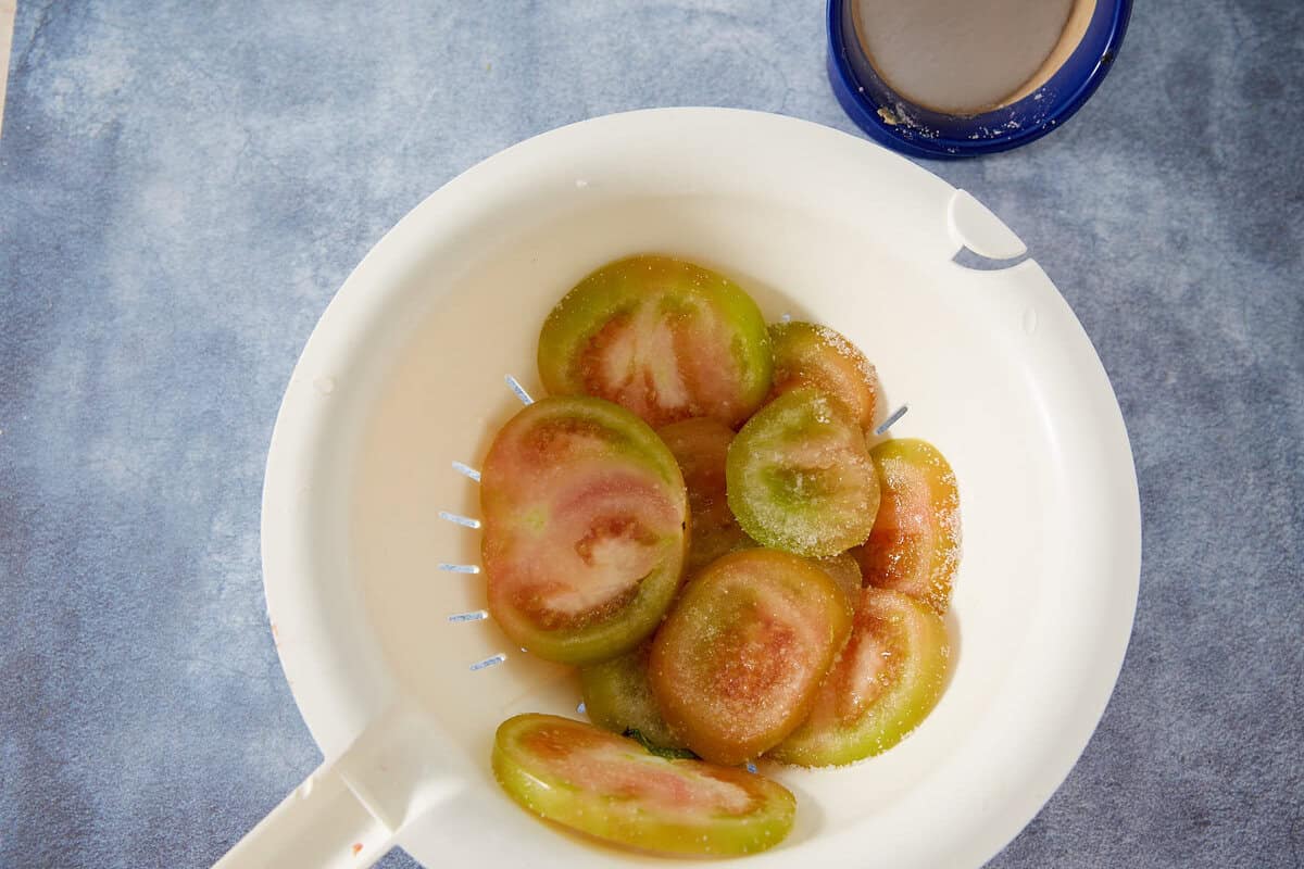 Sliced green tomatoes in a white colander, sprinkled with salt, ready for classic Fried Green Tomatoes, on a blue surface with a blue cup partially visible in the top right corner.