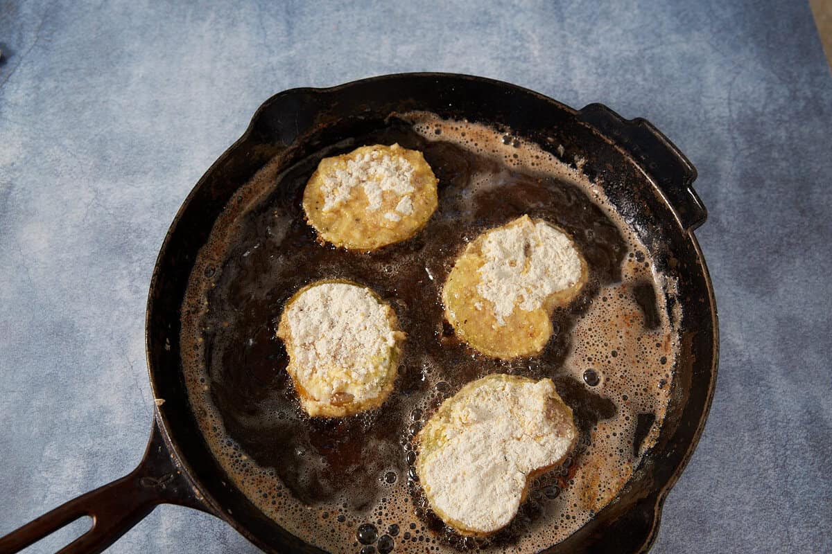 Four battered and flour-coated slices of eggplant are frying in oil in a black cast iron skillet, reminiscent of classic Southern Fried Green Tomatoes, on a light blue textured surface.