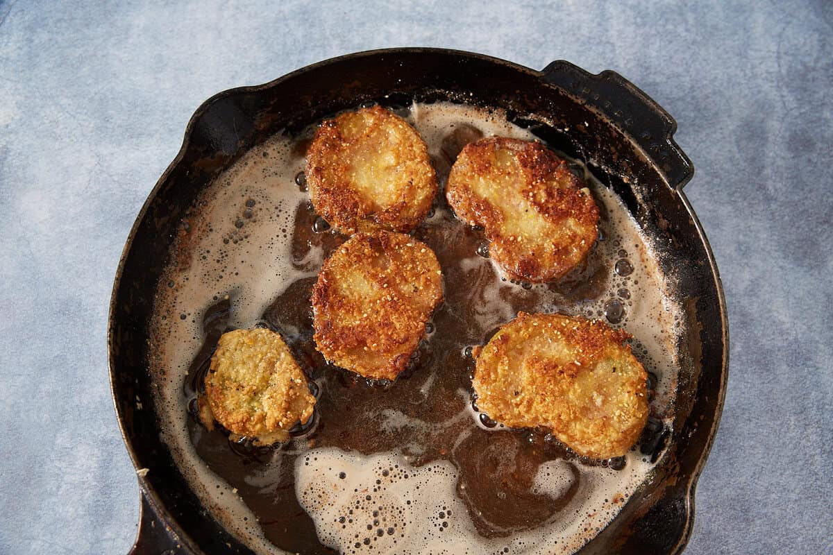 Five golden-brown slices of Fried Green Tomatoes cook in a black cast-iron skillet with bubbling oil, set against a light blue background.