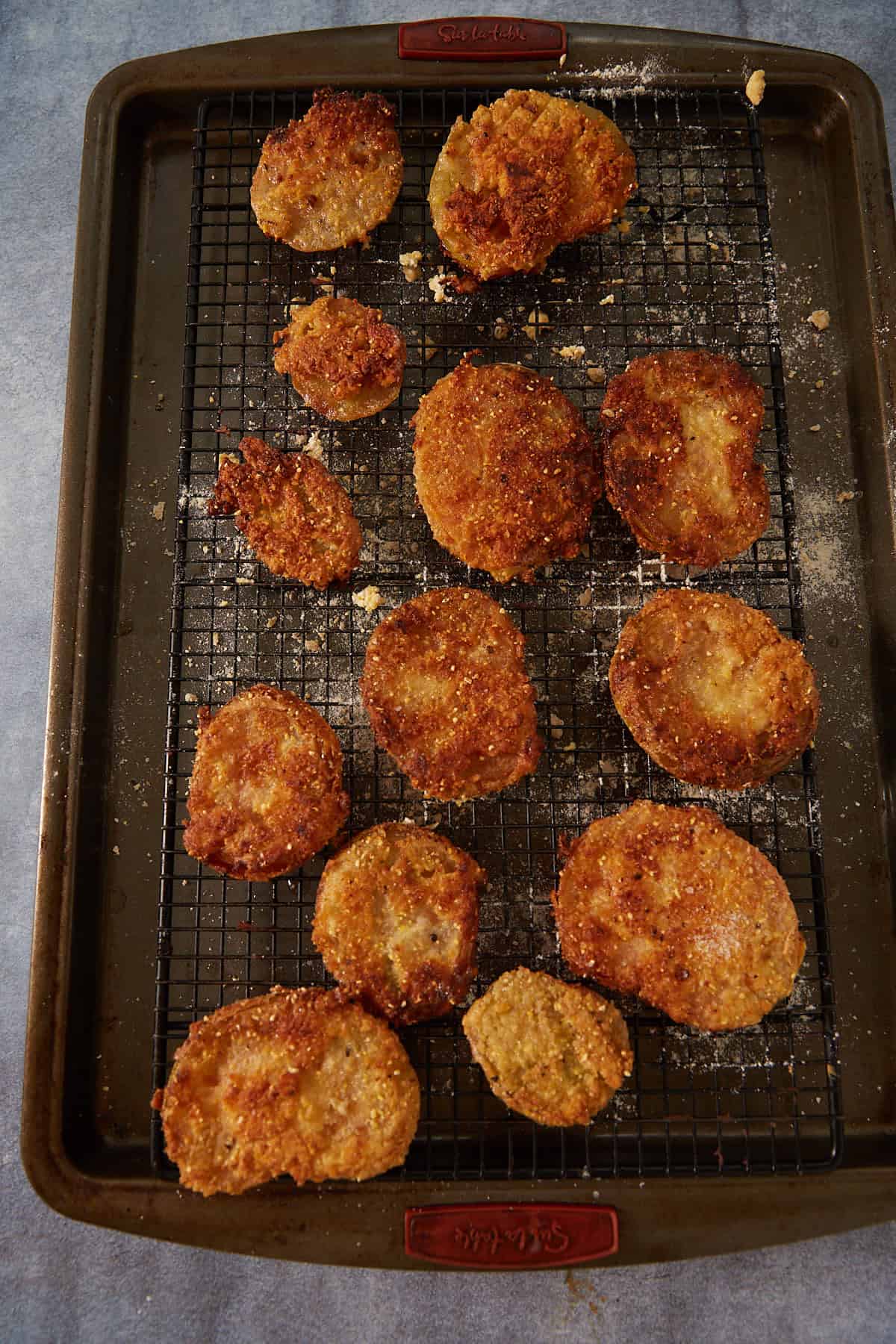 A baking tray with a wire rack holds several pieces of golden-brown fried green tomatoes, sprinkled with crumbs on a blue surface.