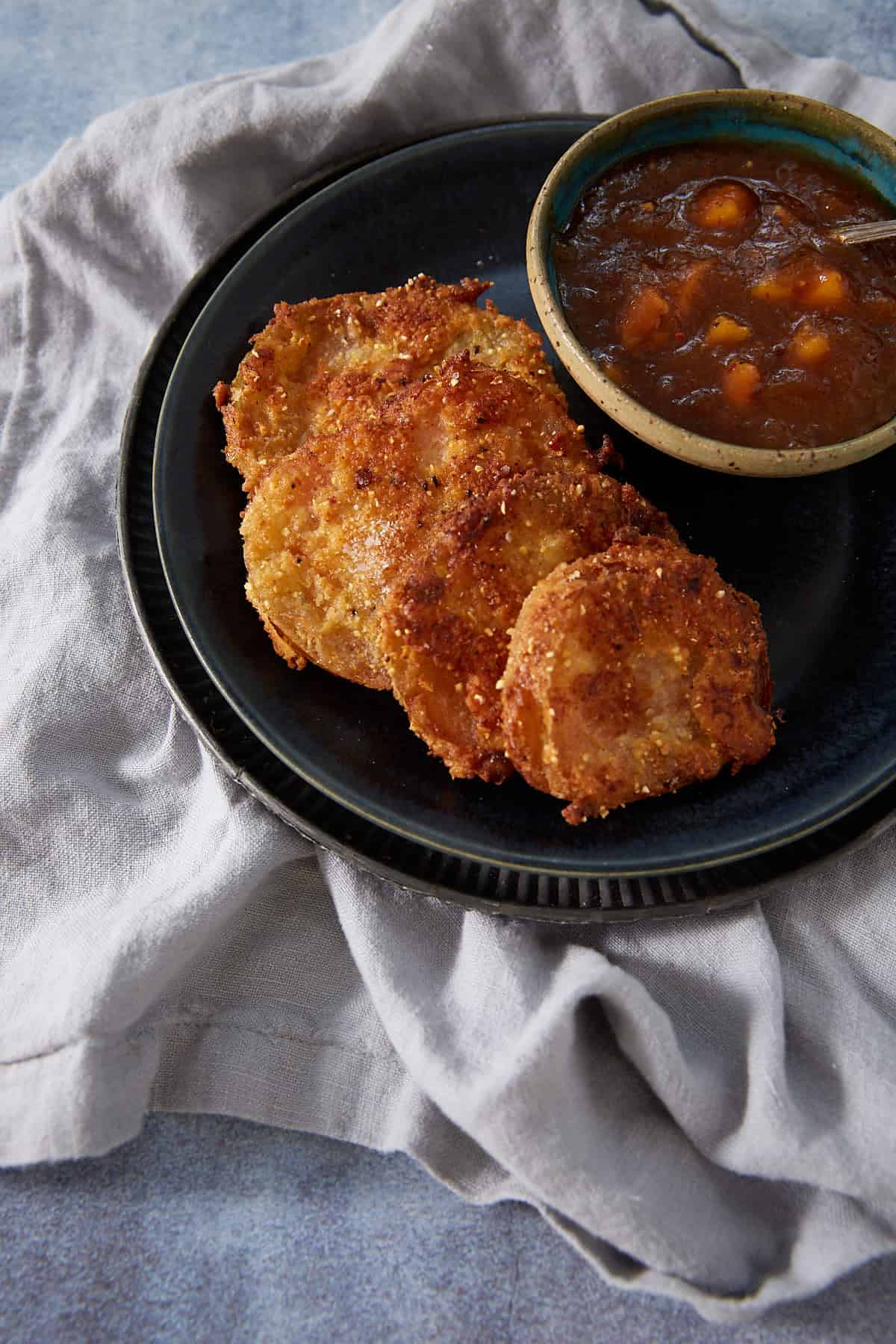 Four crispy, golden-brown Fried Green Tomatoes are arranged on a black plate next to a bowl of chunky brown dipping sauce, all set on a light gray cloth.