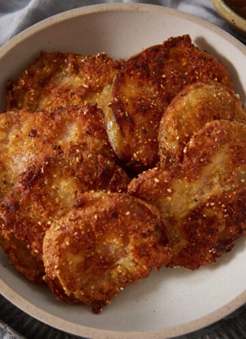 A bowl filled with several golden-brown, crispy fried slices of eggplant, reminiscent of classic Fried Green Tomatoes, arranged in a rustic ceramic dish on top of a dark plate. The slices have a crunchy, textured coating.