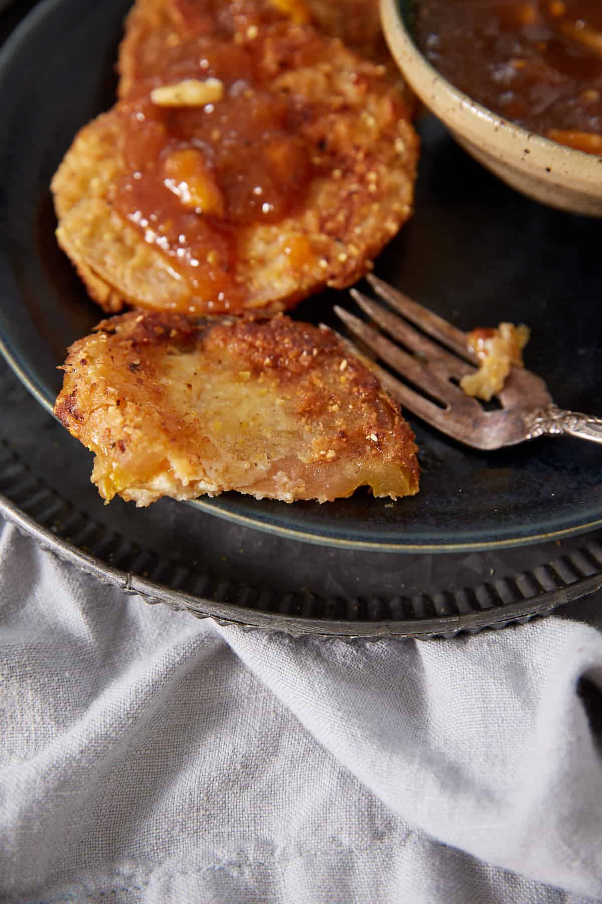 A close-up of Fried Green Tomatoes shows one with a bite taken out, sitting on a dark plate with two whole fried green tomatoes topped with sauce and a fork beside them. A bowl of sauce is partially visible.
