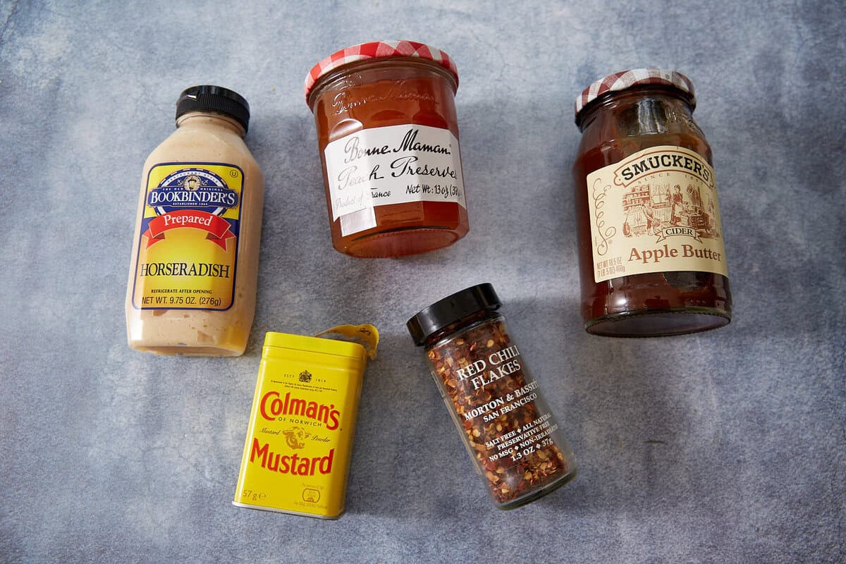 Five condiment jars and containers, including Jezebel Sauce, horseradish, Dijon mustard, red chili flakes, and apple butter, are arranged on a light blue textured surface.