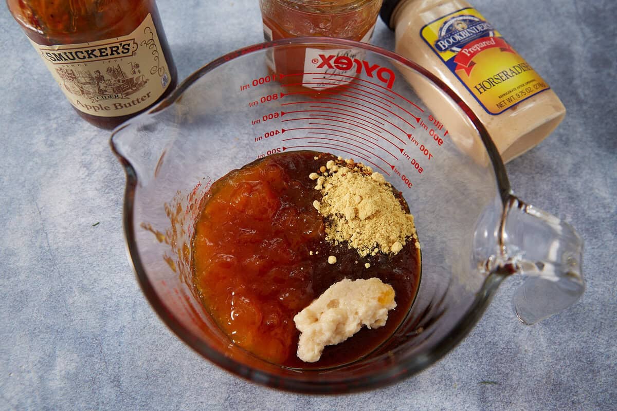 A glass measuring cup contains apple butter, horseradish, ground mustard, and a Jezebel Sauce mixture. Jars of apple butter and horseradish sit nearby on a blue countertop.