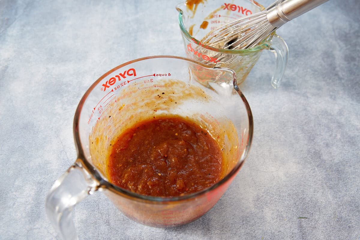 Two Pyrex measuring cups; one in front filled with thick, reddish-brown Jezebel Sauce, and one in the back with sauce residue and a metal whisk inside. Both are on a light gray surface.