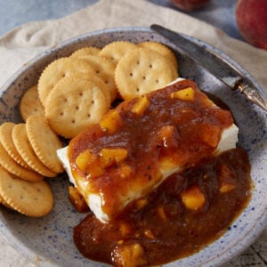 A blue-speckled plate with round crackers and a block of cream cheese topped with chunky peach preserves and a hint of Jezebel Sauce, next to a knife on a beige napkin.