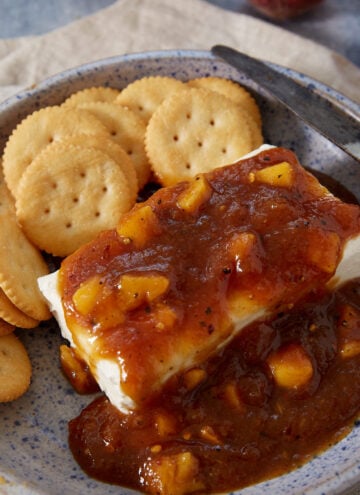 A blue-speckled plate with round crackers and a block of cream cheese topped with chunky peach preserves and a hint of Jezebel Sauce, next to a knife on a beige napkin.