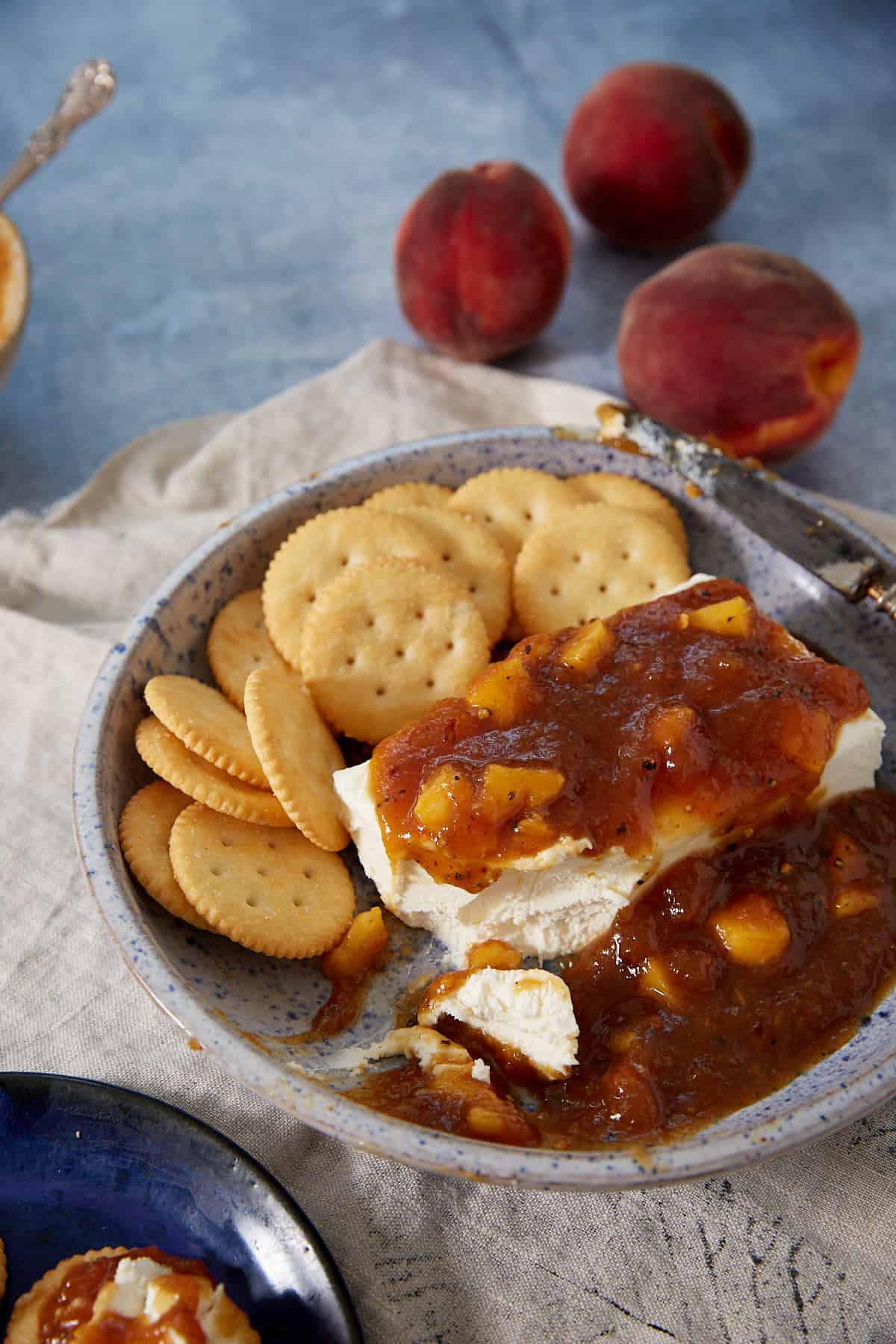 A bowl with round crackers, a block of cream cheese topped with chunky peach Jezebel Sauce, and a knife sits on a linen cloth. Fresh peaches are in the background on a blue surface.