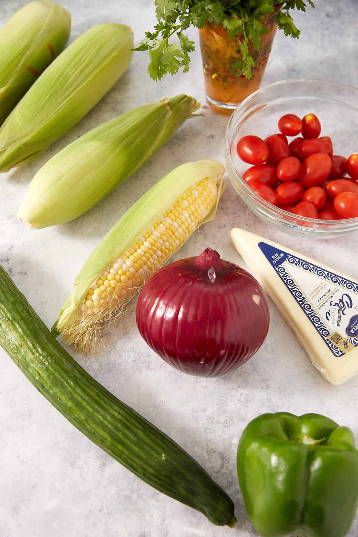 Fresh vegetables including corn on the cob, cherry tomatoes, red onion, cucumber, green bell pepper, a block of cheese, and a bunch of cilantro are arranged on a light countertop.