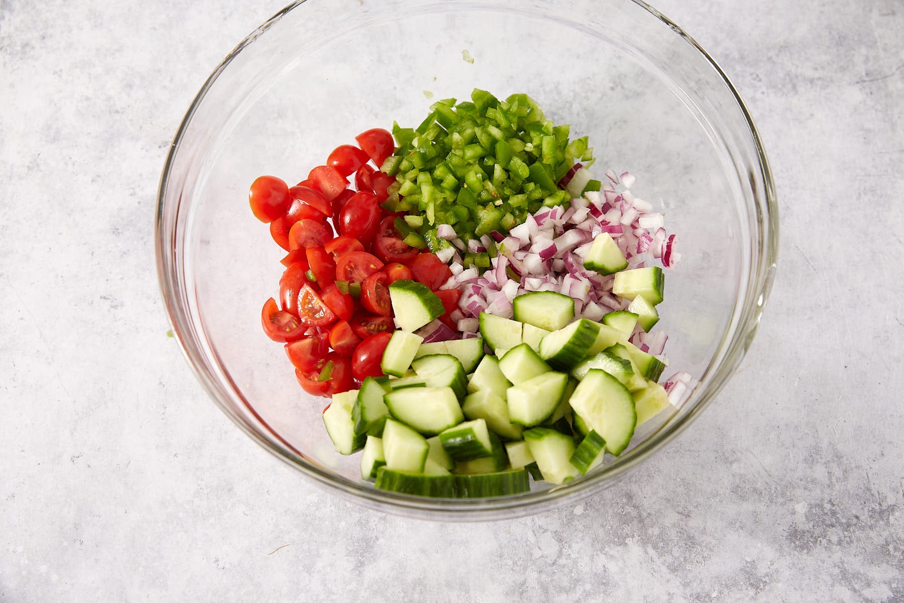 A glass bowl contains chopped cucumbers, halved cherry tomatoes, diced red onion, and diced green bell pepper, arranged in separate sections on a light gray surface.