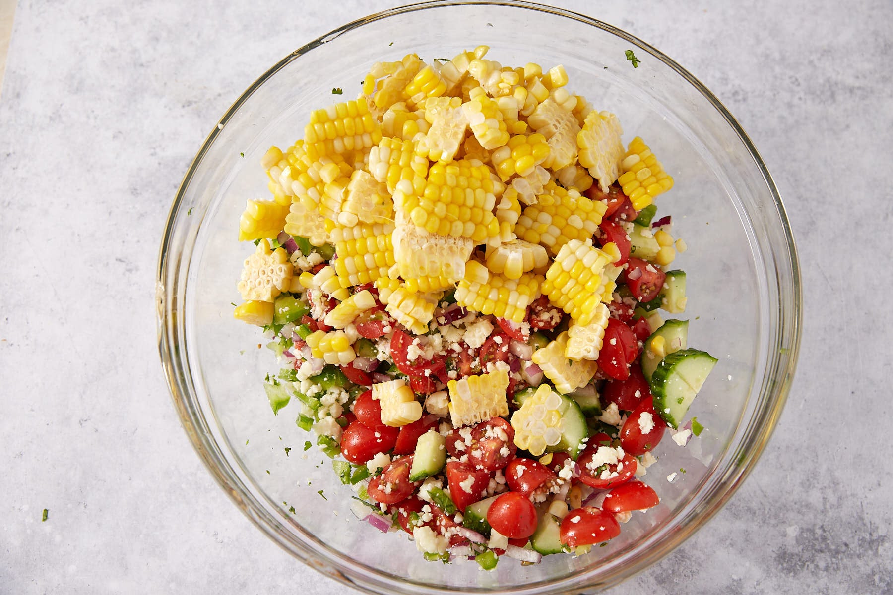 A glass bowl filled with a salad of sliced corn on the cob, cherry tomatoes, diced cucumber, red onion, crumbled cheese, and fresh herbs on a light gray surface.