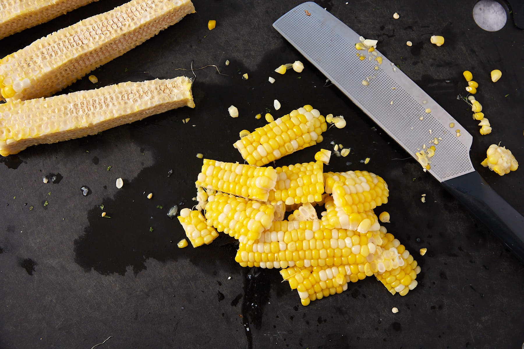 A knife and several pieces of cut corn kernels are on a dark surface next to several empty corn cobs. Corn kernels and bits are scattered around.