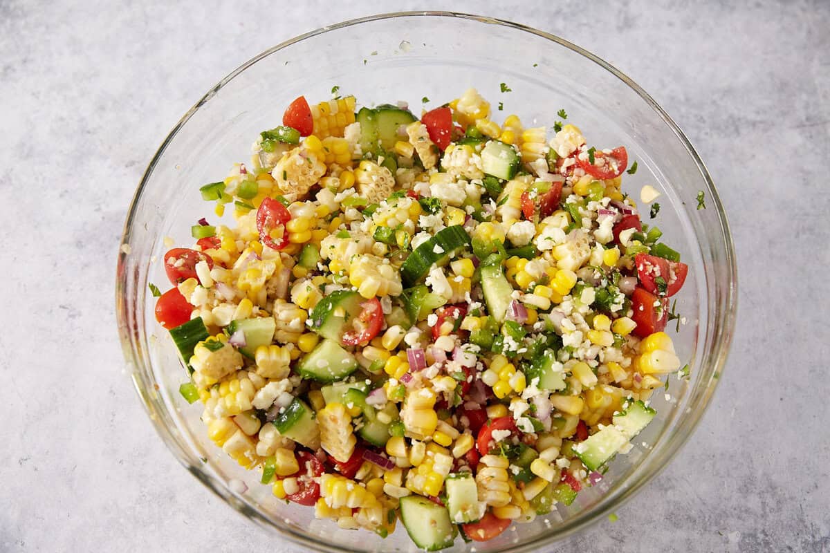 A glass bowl filled with a colorful corn salad, including corn kernels, diced cucumbers, cherry tomatoes, red onions, green peppers, feta cheese, and herbs, on a light gray surface.