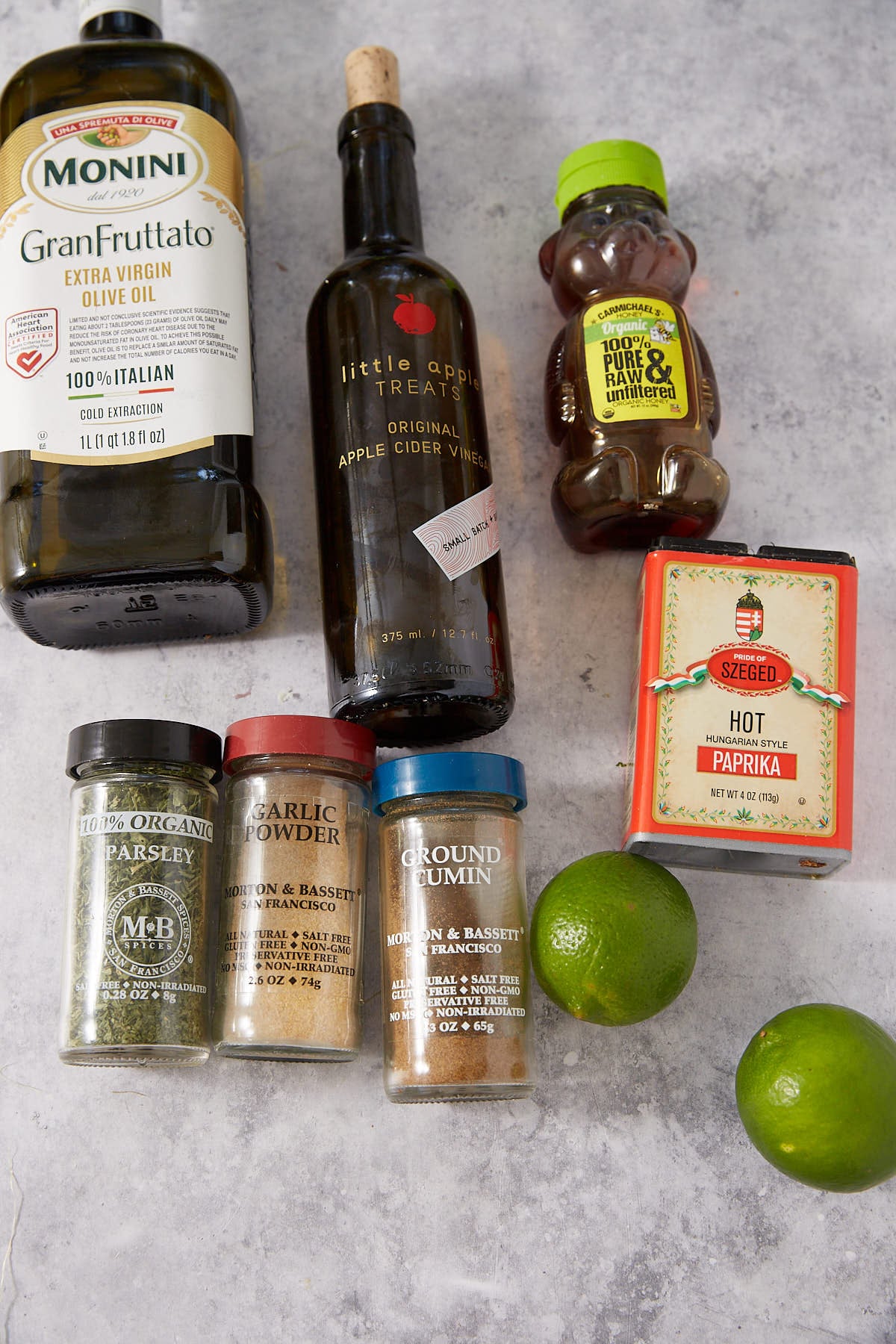 A variety of ingredients on a gray surface, including olive oil, apple cider vinegar, honey, hot paprika, dried parsley, garlic powder, ground cumin, and two whole limes.