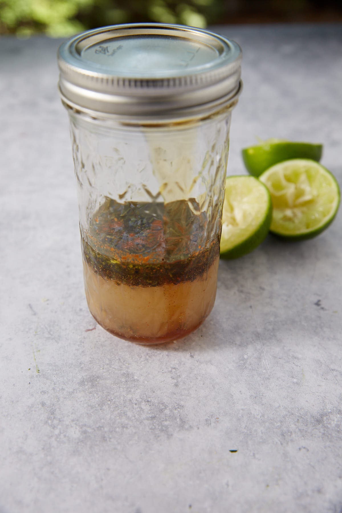 A mason jar filled with a layered liquid mixture sits on a gray surface, with two squeezed lime halves placed beside it.