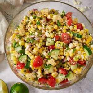 A glass bowl filled with a colorful corn salad containing corn kernels, cherry tomatoes, cucumber, red onion, and herbs. The salad sits on a light surface with a cut lime and a napkin nearby.
