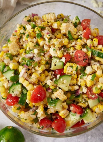A glass bowl filled with a colorful corn salad containing corn kernels, cherry tomatoes, cucumber, red onion, and herbs. The salad sits on a light surface with a cut lime and a napkin nearby.