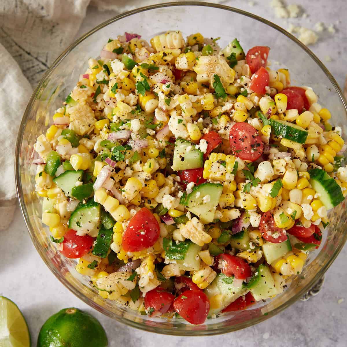 A glass bowl filled with a colorful corn salad containing corn kernels, cherry tomatoes, cucumber, red onion, and herbs. The salad sits on a light surface with a cut lime and a napkin nearby.
