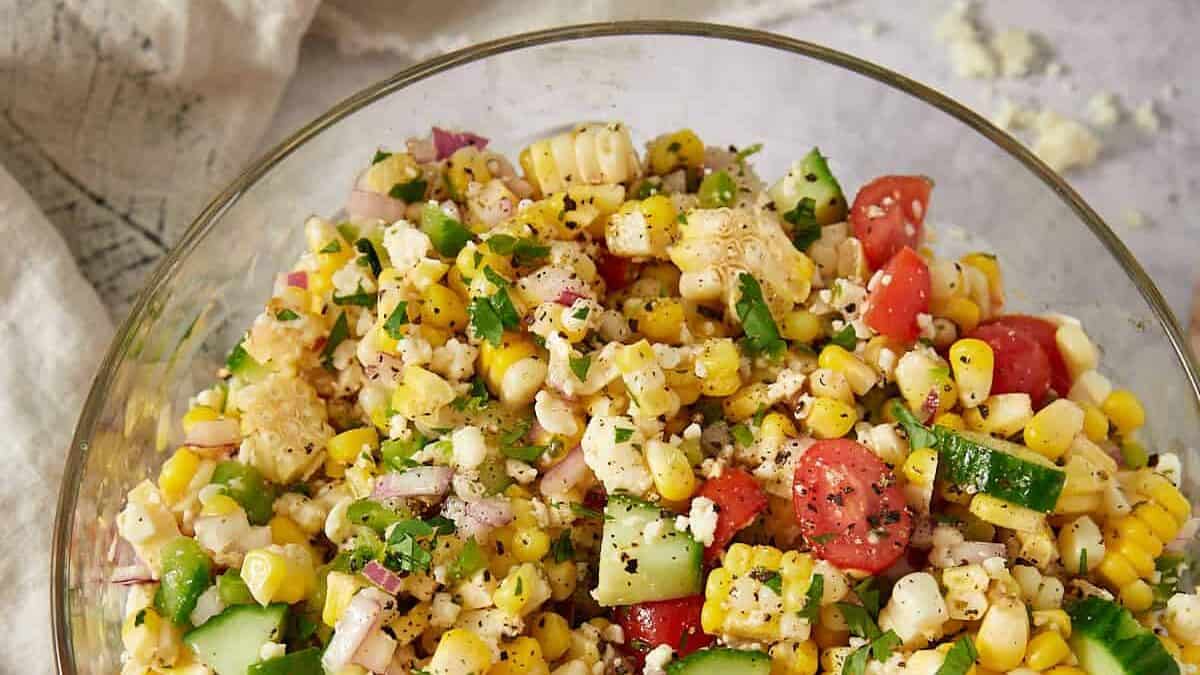 A glass bowl filled with a colorful corn salad containing corn, cherry tomatoes, cucumbers