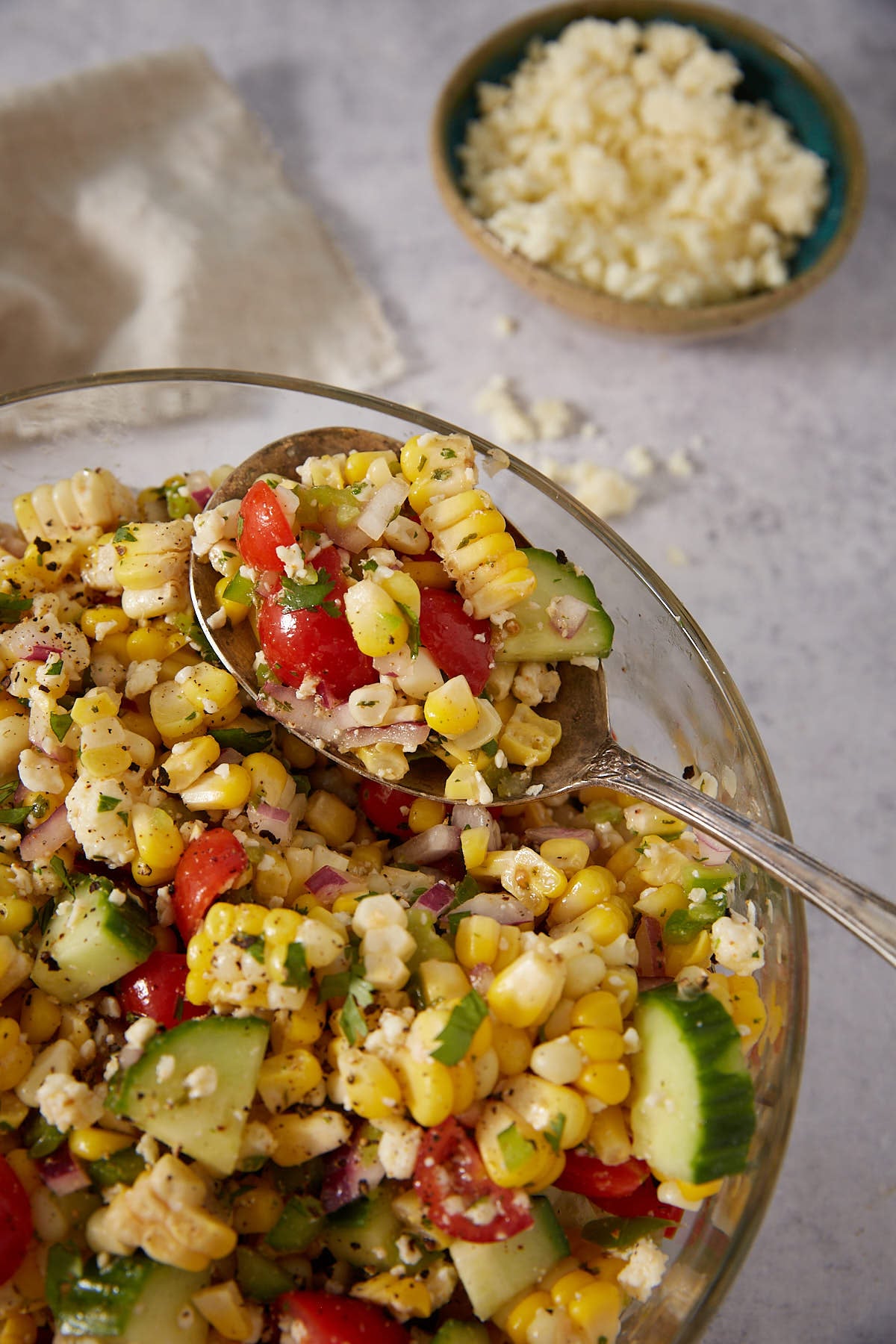 A glass bowl of fresh corn salad with cucumbers, tomatoes, red onion, herbs, and crumbled cheese, with a spoon inside. A small dish of more crumbled cheese is in the background.