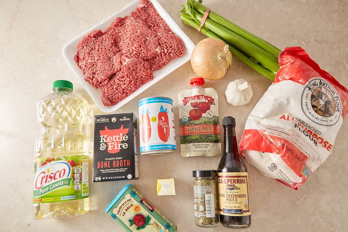 A countertop displays ground beef, celery, onion, garlic, all-purpose flour, canola oil, beef broth, canned tomatoes, tomato paste, Worcestershire sauce, dried herbs, bay leaf, and a small cube of butter.